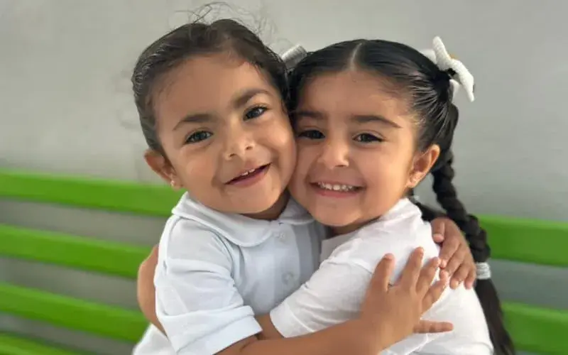 Dos estudiantes sonrientes se abrazan frente a una pared con franja verde en el colegio