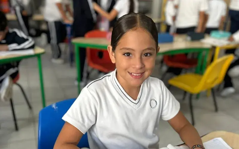 Estudiante sonriendo en el aula durante clase en el instituto, rodeada de compañeros y mobiliario escolar colorido